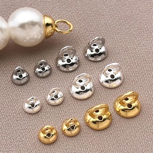 May include: A selection of small, metallic jewellery findings in silver and gold. The findings are bell-shaped with a loop at the top and a hole for attachment. A large pearl is also visible in the image.