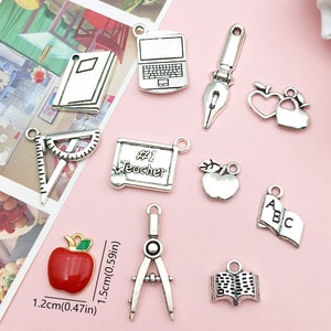 May include: A set of 10 silver charm pendants with school-themed designs, including an apple, a book, a laptop, a compass, a ruler, a paintbrush, a heart, and a chalkboard with the text "#1 Teacher".