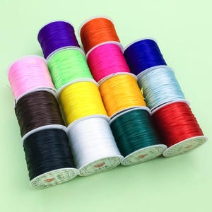 Puede incluir: Una colección de coloridas bobinas de cordón elástico en varios tonos, incluyendo rosa, verde, amarillo y azul. Las bobinas están cuidadosamente dispuestas, mostrando los colores vibrantes de los cordones. Los cordones se utilizan probablemente para hacer joyas o manualidades.