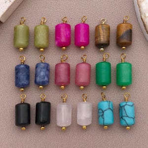 May include: A collection of cylindrical gemstone beads in various colors, including green, pink, blue, and brown. Each bead has a gold-colored loop at the top and a gold-colored bead at the bottom, ready for jewelry making. The beads are arranged on a neutral background.