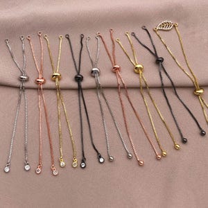 May include: A collection of adjustable bracelet chains in various metallic finishes, including silver, gold, rose gold, and black. Each chain features a sliding bead for adjustment and a small charm or bead at the end. A leaf-shaped charm is also included.