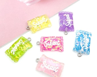 Pode incluir: Seis encantos coloridos em forma de doce com argolas de salto prateadas. Cada encanto é translúcido e apresenta uma inscrição "Sweet" em branco. Os encantos são em tons de rosa, roxo, azul, verde e amarelo.