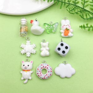 May include: A set of 10 white and black charm pendants, including a snowflake, a dice, a doughnut, a cloud, a cat, a bottle, a butterfly, a duck, a gummy bear, and a bunny holding a carrot. The charms are made of polymer clay and have a silver-toned metal loop for attaching to a necklace or bracelet.