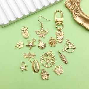 May include: A collection of gold-toned metal charms featuring various designs, including owls, butterflies, seashells, stars, trees, and a dragonfly. The charms are arranged on a green surface.