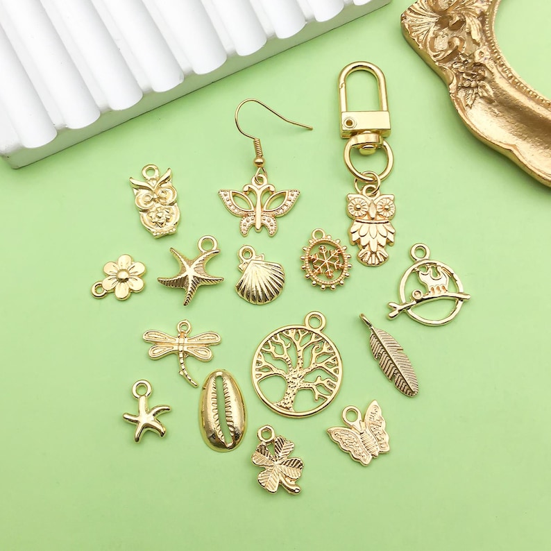 May include: A collection of gold-toned metal charms featuring various designs, including owls, butterflies, seashells, stars, trees, and a dragonfly. The charms are arranged on a green surface.