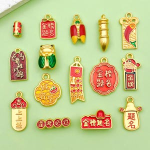 May include: A collection of gold-toned metal charms featuring colorful enamel accents. The charms include various shapes such as rectangular tags, a frog, a fish, a brush, and circular designs, all decorated with Chinese characters and vibrant red, yellow, and green patterns.