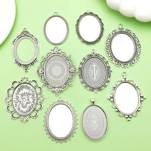 May include: A selection of silver-coloured oval pendant trays with decorative, ornate frames. Some have clear inserts, while others have textured or patterned surfaces. The pendants are arranged on a light green surface.