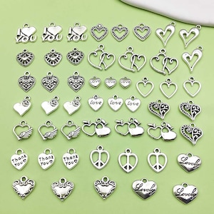 Op de afbeelding: Een verzameling zilverkleurige metalen bedels in de vorm van harten, vleugels en vredestekens. Sommige bedels hebben de tekst "Love", "Thank You" en "Lovely".