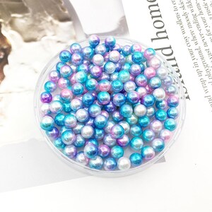 May include: A clear plastic container filled with iridescent blue, pink, and white pearl beads. The beads are small and round, and they have a shimmery finish.