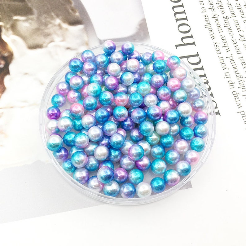 May include: A clear plastic container filled with iridescent blue, pink, and white pearl beads. The beads are small and round, and they have a shimmery finish.