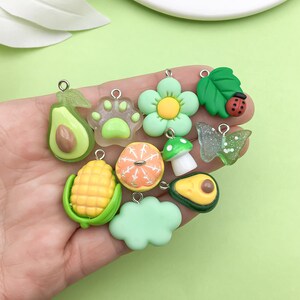 Puede incluir: Un conjunto de 10 colgantes de charms verdes, amarillos y naranjas con diversas formas, incluyendo una huella de pata, un aguacate, ma&iacute;z, un hongo, una flor y una mariquita. Los charms est&aacute;n hechos de arcilla polim&eacute;rica y tienen un acabado brillante.