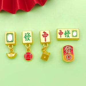 May include: Five gold-colored charms with Chinese characters and symbols. The charms are in the shape of mahjong tiles and feature a gold coin, a red circle, a dice, and a gold bar with the Chinese character for "luck".