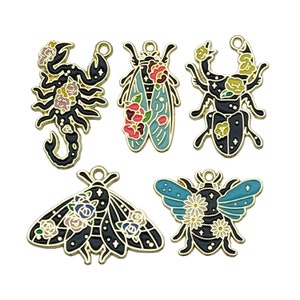 Pode incluir: Seis pingentes de metal dourado com desenhos de insetos e escorpiões em preto e branco com detalhes florais coloridos. Os pingentes têm um laço na parte superior para serem presos a joias ou outros trabalhos manuais.