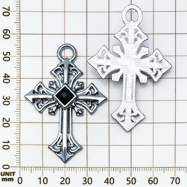 May include: Two silver-colored crosses. One cross features a black square gem at the center. Both crosses have ornate, gothic-style designs with a loop at the top for hanging. The crosses are displayed against a grid background.