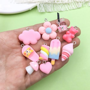 May include: A set of 10 pink and white charm pendants featuring various cute food and drink designs, including a cloud, a flower, a popsicle, a marshmallow, a slipper, a mushroom, a heart, a bear, a cone, and a glass of juice.