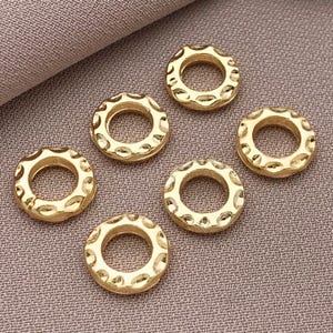 Pu&ograve; includere: Sei anelli metallici color oro dall'aspetto martellato con un design circolare. Gli anelli hanno una superficie strutturata e un foro al centro. Gli anelli sono disposti su un tessuto di colore neutro.