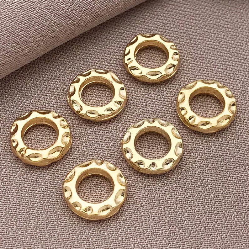 Pu&ograve; includere: Sei anelli metallici color oro dall'aspetto martellato con un design circolare. Gli anelli hanno una superficie strutturata e un foro al centro. Gli anelli sono disposti su un tessuto di colore neutro.