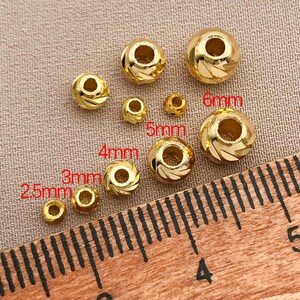 May include: Assortment of gold-coloured beads, sizes from 2.5mm to 6mm. Some beads feature a spiral pattern, others are plain. A ruler is present for scale. The beads are measured in millimetres.