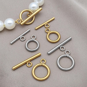 Könnte beinhalten: Nahaufnahme von gold- und silberfarbenen Knebelverschlüssen. Die Verschlüsse haben ein Stangen- und Ringdesign in verschiedenen Größen. Einige sind an einer Perlenkette befestigt. Die Verschlüsse sind auf einem neutralfarbenen Stoff abgebildet.