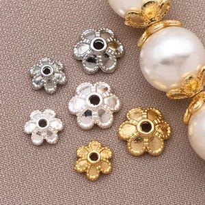 May include: Close-up of several flower-shaped jewelry components in silver and gold tones. The components have a central hole and are arranged on a textured, neutral-colored surface. Some are paired with large, white, pearl-like beads.