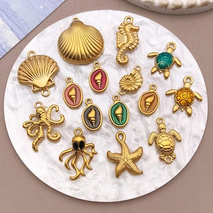 May include: A selection of gold-coloured sea-themed charms, including seashells, seahorses, turtles, octopuses, and starfish. Some charms feature colourful enamel details. The charms are arranged on a white, marbled surface.