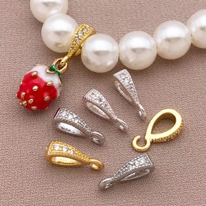 May include: A collection of jewelry findings, including gold and silver-toned bails and a strawberry charm. The bails are adorned with clear crystals. A pearl necklace is in the background.
