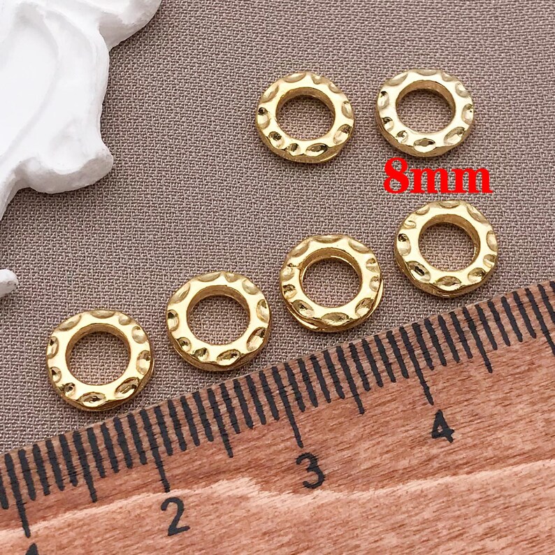 Pu&ograve; includere: Sei perline circolari dorate e strutturate, ciascuna di circa 8 mm di diametro, sono disposte su una superficie marrone. Un righello di legno con segni neri &egrave; in primo piano. Le perline hanno un motivo decorativo sul bordo esterno.