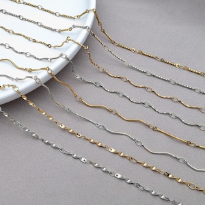 Puede incluir: Una variedad de delicados collares en tonos dorados y plateados. Las cadenas presentan varios diseños, incluyendo eslabones ovalados, patrones retorcidos y líneas onduladas. Las joyas se exhiben sobre un plato blanco y una superficie gris.
