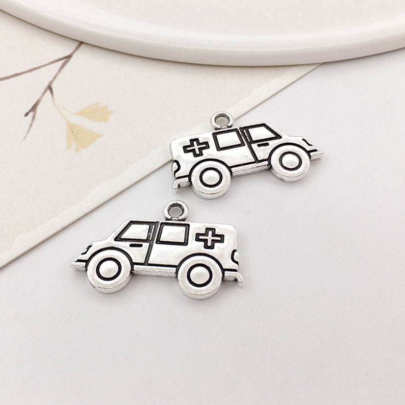 May include: Two silver-toned metal charms shaped like ambulances. Each charm has a black outline and a black cross on the side.
