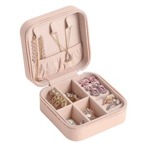 May include: A pale pink, square-shaped jewelry box with a zipper closure. The box is open and contains various jewelry pieces, including necklaces, earrings, and rings.