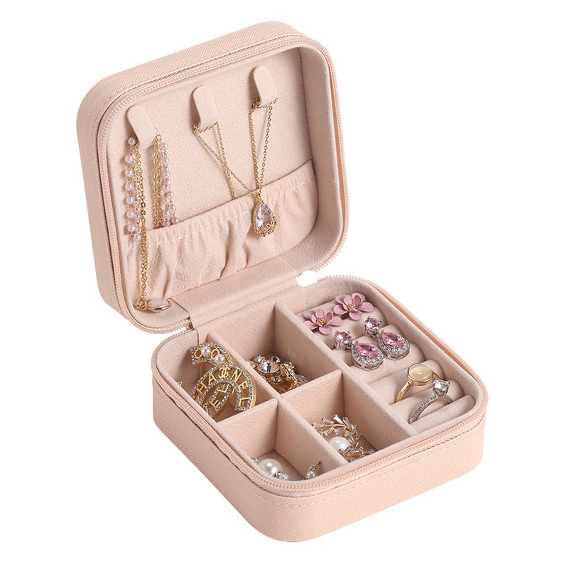 May include: A pale pink, square-shaped jewelry box with a zipper closure. The box is open and contains various jewelry pieces, including necklaces, earrings, and rings.