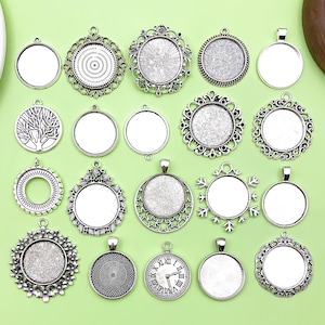 Peut inclure: Une collection de pendentifs argentés de différentes formes et designs. Les plateaux comprennent des formes rondes, ovales et en forme de flocon de neige, avec des filigranes décoratifs et des centres vierges pour la personnalisation. Les pendentifs sont présentés sur une surface verte.