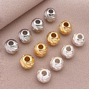 May include: A selection of silver and gold-coloured round beads with a textured, etched design. The beads have a hole through the centre, and are scattered on a pink fabric surface. These beads are suitable for jewellery making.
