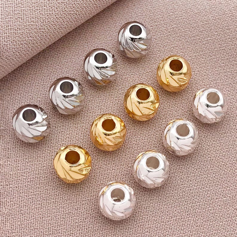 May include: A selection of silver and gold-coloured round beads with a textured, etched design. The beads have a hole through the centre, and are scattered on a pink fabric surface. These beads are suitable for jewellery making.