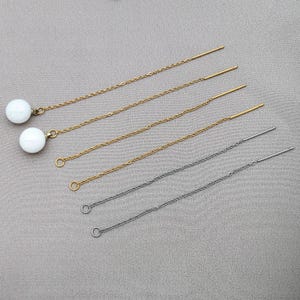 May include: A collection of delicate threader earrings. Two pairs feature a gold chain with a white, round bead. The other two pairs have a gold chain and a silver chain, respectively. The earrings are displayed on a light gray surface.