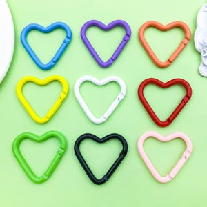 May include: Nine heart-shaped carabiner clips in a range of colours: blue, purple, orange, yellow, white, red, green, black, and pink. Each clip has a spring-loaded gate for secure attachment. They are arranged on a light green surface.