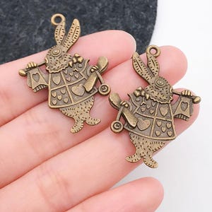 May include: Two antique gold-toned metal charms of a rabbit holding a playing card and riding a toy scooter. The rabbit is wearing a vest with a heart pattern.