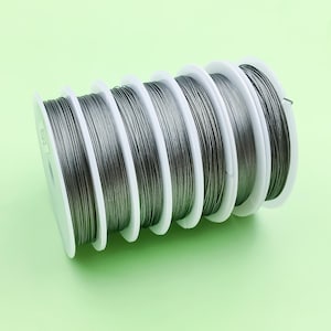 May include: A series of spools, each holding grey wire, are displayed. The wire is wound around white plastic spools, creating a neat, organised arrangement. The metallic wire contrasts with the white spools, set against a light green backdrop. Suitable for crafts or jewellery.