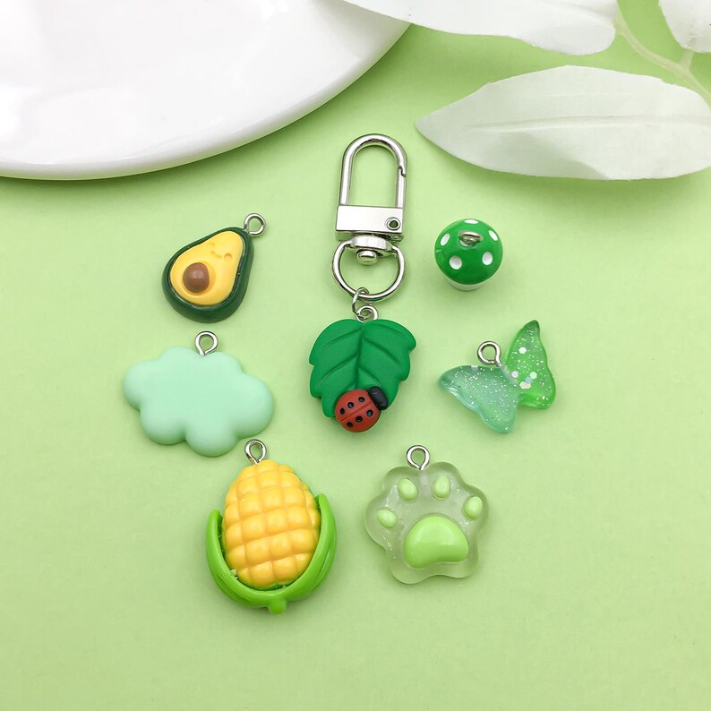 Puede incluir: Un conjunto de siete dijes verdes y amarillos con un llavero plateado. Los dijes incluyen un hongo, una hoja con una mariquita, una nube, un aguacate, ma&iacute;z, una huella de pata y una mariposa.