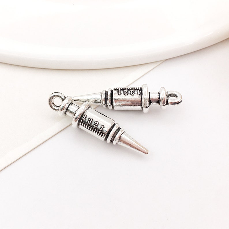 May include: Two silver-toned metal syringe charms with black markings. The charms are in the shape of a syringe with a needle and a plunger. The markings on the syringes read "4321" and "4321".