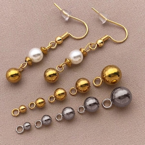 Può includere: Componenti di gioielleria color oro e argento, tra cui orecchini e perline rotonde di varie dimensioni. Gli orecchini presentano ganci, catene e perline color oro, con un accento di perla bianca. Sono presenti anche perline dorate e argentate aggiuntive.