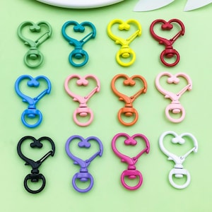 Puede incluir: Una colección de doce cierres giratorios en forma de corazón en varios colores, incluyendo verde, turquesa, amarillo, rojo, azul, rosa, naranja, negro, morado, magenta y blanco. Cada cierre tiene un gancho giratorio de metal y un anillo circular.