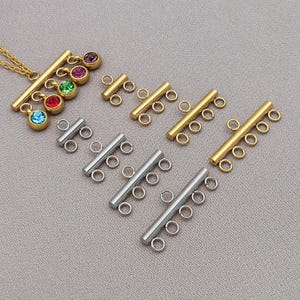 Può includere: Componenti per gioielli dorati e argentati. I pezzi dorati includono una collana con ciondoli di pietre preziose colorate e più connettori a barra. I pezzi argentati sono connettori a barra. Tutti i pezzi hanno piccoli anelli per il fissaggio.