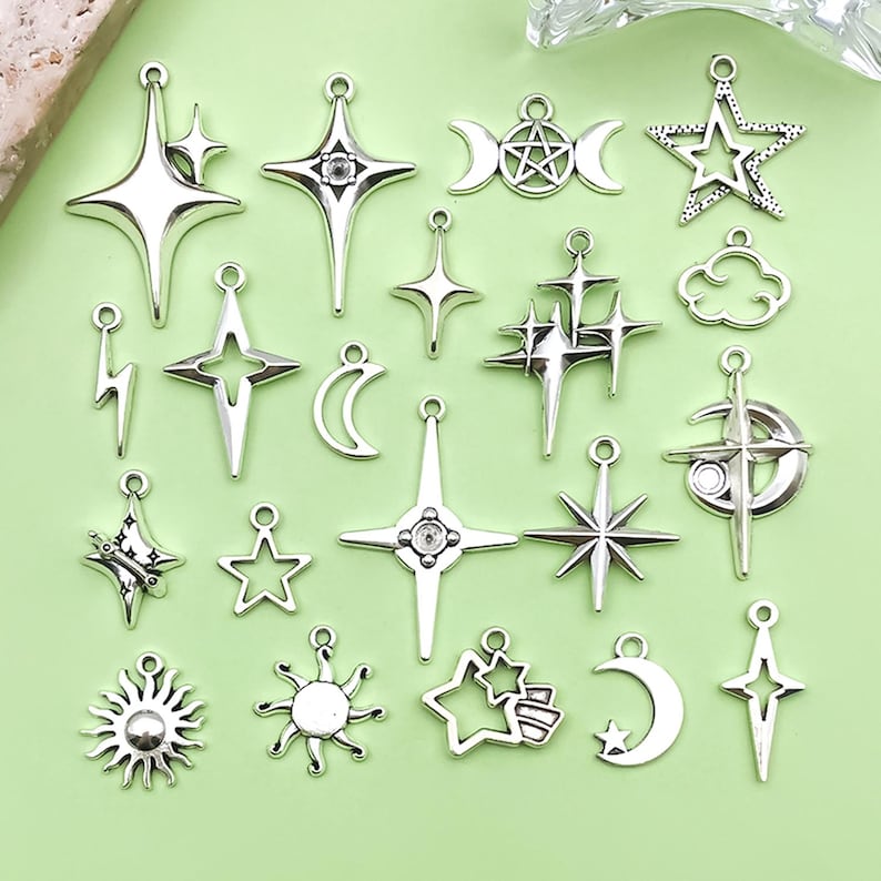 K&ouml;nnte beinhalten: Eine Sammlung silberfarbener Anh&auml;nger in verschiedenen Himmelsk&ouml;rper-Formen, darunter Sterne, Monde, Sonnen und Kreuze. Die Charms haben eine metallische Oberfl&auml;che und sind mit aufwendigen Details versehen, ideal f&uuml;r Schmuckherstellung oder Bastelprojekte.