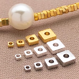 May include: Various gold and silver square beads with a central hole, presented with a gold chain and a large white pearl. The beads are of different sizes, suitable for jewellery making.