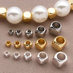 May include: An assortment of jewelry beads in various sizes and finishes. The beads include large, round, white faux pearls, gold-colored cube beads, and silver and gold-colored cube-shaped beads with a hole through the center. The beads are arranged on a textured, tan surface.