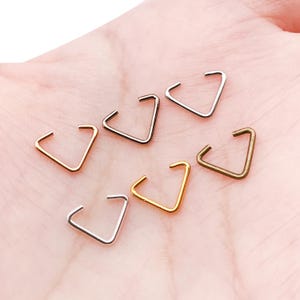 以下が含まれることがあります： 金、銀、ブロンズなど、さまざまな金属仕上げの三角形のイヤーカフ7個セット。 イヤーカフは、幾何学的なデザインと多様なカラーパレットを示す、明るいピンク色の表面に配置されています。
