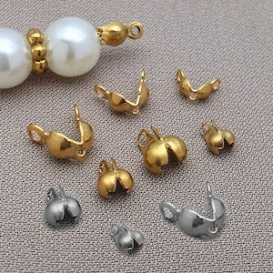 Könnte beinhalten: Eine Sammlung gold- und silberfarbener Schmuckzubehörteile, darunter Perlkappen und Verschlüsse. Die Fundstücke sind in verschiedenen Formen und Größen gehalten, mit einer Perlenkette, die teilweise in der oberen linken Ecke zu sehen ist. Der Hintergrund ist ein neutrales Grau.