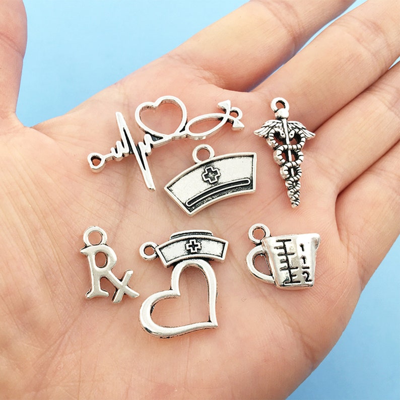 May include: A collection of silver charm pendants with medical and nursing themes. The charms include a heart, a stethoscope, a nurse's cap, a measuring cup, and a caduceus.