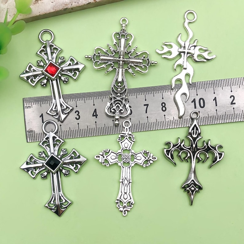 May include: Six silver-toned cross pendants with intricate designs. One cross features a red gem, another a black gem. A ruler shows measurements in inches and centimeters, indicating the size of the crosses. The crosses have various gothic and ornate styles.